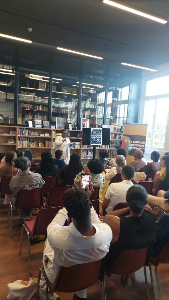 A speaker presenting at an event in a library, with an audience seated and engaged. The setting features bookshelves and a banner for a conference titled 'Black Scholar and Expert Conference.'