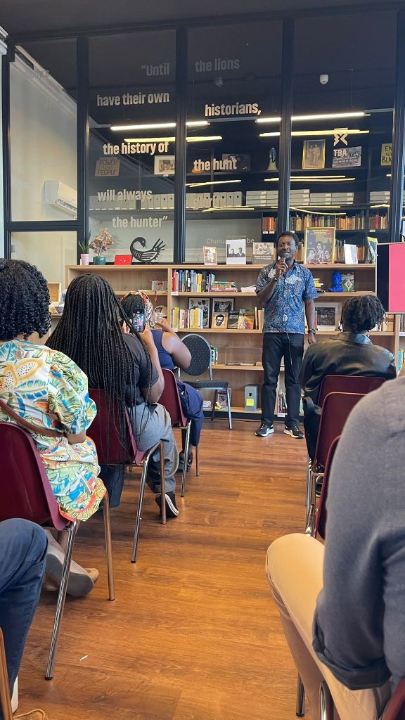 A speaker addresses an audience in a library setting, featuring bookshelves in the background with a motivational quote on the wall.