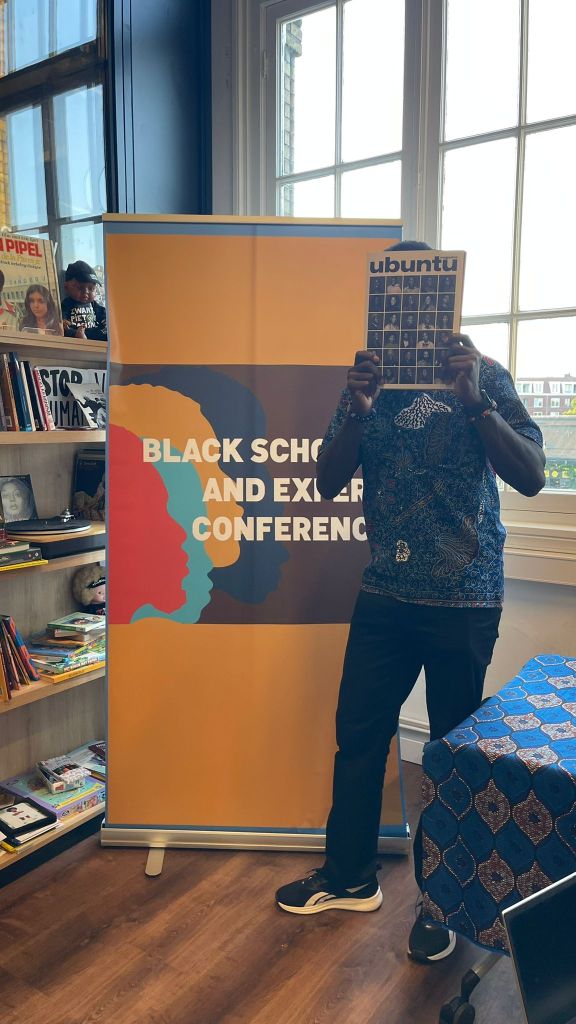 A person holding a magazine titled 'Ubuntu' in front of a banner that reads 'Black Scholars and Excellence Conference'. The background features a bookshelf with various items visible.
