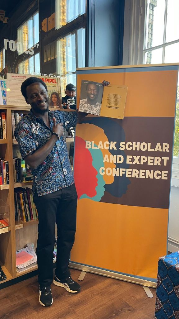 A smiling individual stands next to a banner for the 'Black Scholar and Expert Conference' in a book-filled environment, pointing towards an image on the banner.